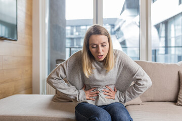 Young woman sitting on sofa holding hands on stomach twisted from pain, poisoned or menstrual pain, woman at home