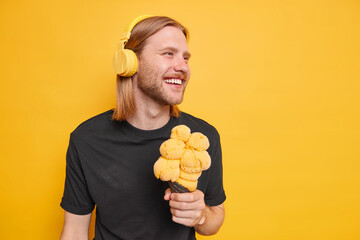 Handsome cheerful man with long red hair looks aside happily wears wireless headphones listens music eats appetizing ice cream dressed casually isolated over yellow background blank free space