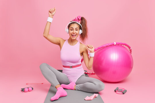 Cheerful female athlete with pony tail in activewear enjoys favorite playlist via wireless headphones poses on fitness mat has training at home surrounded by sport equipment isolated on pink wall