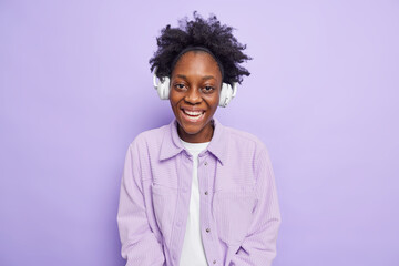 Fototapeta na wymiar Cheerful dark skinned teenage girl smiles happily enjoys listening music smiles toothily uses wireless headphones dressed casually isolated over purple background. Youth emotions hobby concept