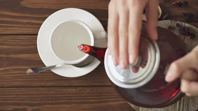 The process of brewing red tea with hibiscus. Step four. Female hands pouring fruity red tea into a white mug. Textured wood kitchen table, wood cut and glass teapot with hot tea. Dry hibiscus.