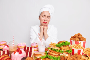 Binge eating concept. Stressed unhappy Asian lady wants to eat fast food tempting and looks with appetite has addiction to cheat meal wears comfortable bathrobe towel over head poses indoor.
