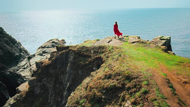 Strong wind blowing a red dress of a young woman standing by the edge of a cliff. A beautiful girl enjoying rocky seashore and scenic landscapes with flying birds in Lizard, Cornwall, England in 4K.