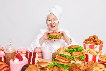 Time for snack. Happy old lady eats appetizing hamburger smiles with cheerful expression applies beauty patches under eyes feels temptation to eat high calorie food. Unhealthy fastfood addiction