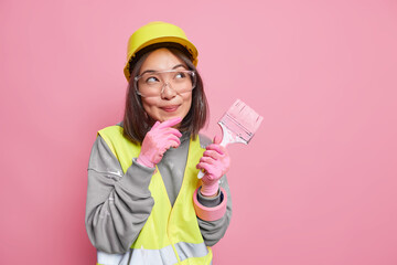 Dreamy female building contractor makes plans thinks about new decor of apartment going to paint walls wears safety glasses helmet uniform holds painting brush isolated over pink background.