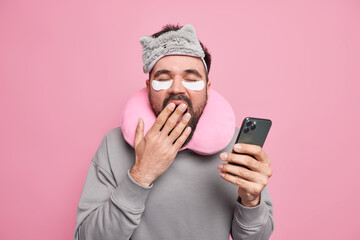 Photo of sleepy man covers mouth with hand wants to have rest scrolls social networks via smartphone applies patches to reduce puffiness under eyes wears sleepmask travel pillow around neck.
