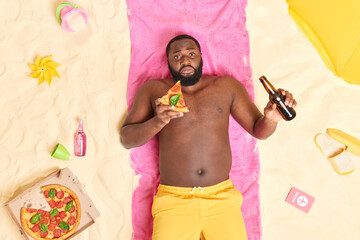 Outdoor shot of unhappy bearded Afro American man eats pizza and drinks beer enjoys good rest at...