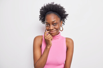 Fototapeta na wymiar Portrait of good looking dark skinned woman with curly hair smiles gently keeps finger near lips wears earrings pink t shirt isolated over white background. Pleasant face expressions and feelings