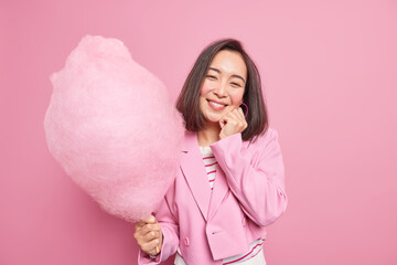 Beautiful brunette young woman smiles gently keeps hand on face holds candy floss on stick wears...