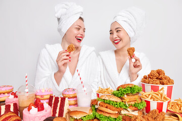 Two happy diverse women look gladfully at each other hold nuggets eat tasty fast food enjoy cheat meal dressed in casual domestic clothes isolated over white background. Unhealthy nutrition.