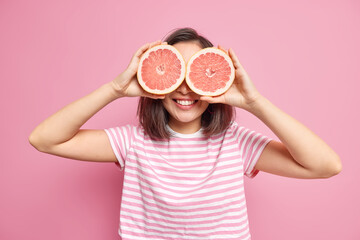 Happy brunette woman covers eyes with fresh juicy grapefruit slices smiles gladfully keeps to...
