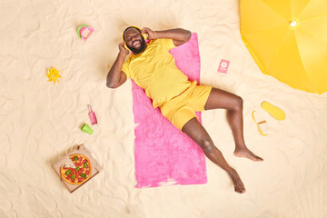 Obraz na płótnie Canvas Happy dark skinned man dressed in yellow summer costume listens music via headphones eats fast food spends lazy summer day at beach has overjoyed expression. Recreation lifestyle travel concept