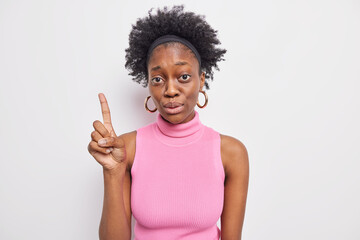 Surprised dark skinned Afro American woman with natural curly hair points index finger up shows something amazing wears pink t shirt earrings isolated over white background. Just look there.
