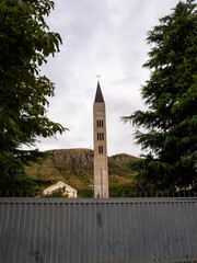Tower of the Catholic Church and Franciscan Monastery framed by a roof, trees and mountain.
Mostar,...