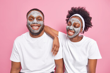 Glad Afro American couple apply clay facial mask for face care smile happily have good mood dressed in casual white t shirt spend free time together isolated over pink background. Beauty treatment - Powered by Adobe