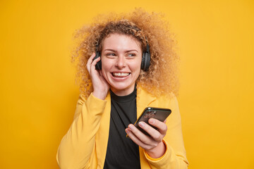 Fototapeta na wymiar Image of cheerful curly woman uses modern technologies for listening music smiles toothily holds mobile phone wireless headphones on ears poses against yellow background. People leisure concept
