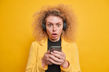 Stupefied young woman with natural curly hair uses mobile phone for listening music wears wireless headphones on ears dressed in formal clothes isolated over yellow background. Human reactions