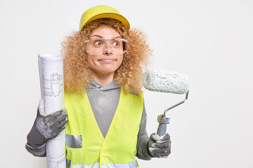 Surprised curly haired woman presses lips focused into distance wears protective helmet uniform and...