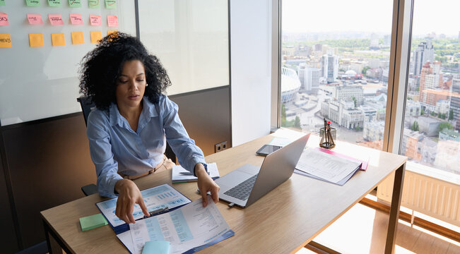 African American businesswoman ceo analyst sitting at desk working with papers with laptop near panoramic window in contemporary corporation office. Financial data analyst working with big data. - Powered by Adobe