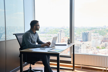 Indian latin successful thoughtful businessman leader thinking of future corporate successful plan ideas sitting at office desk looking at panoramic view window of building of modern corporation. - obrazy, fototapety, plakaty