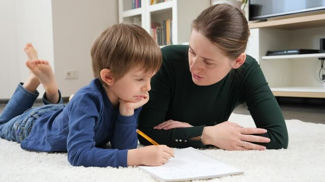 Angry mother scolding and talking to her silly son doing homework on floor in living room. Concept of domestic education, child development and parenting problems