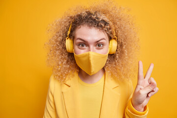 Cheerful curly haired Caucasian woman makes peace gesture enjoys free time listens music via headphones wears protective face mask during coronavirus outbreak isolated over yellow background.