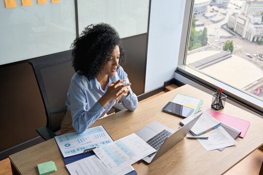 Young African American businesswoman ceo sitting at desk having videocall on yearly financial report with colleagues using laptop near panoramic window in contemporary corporation office.