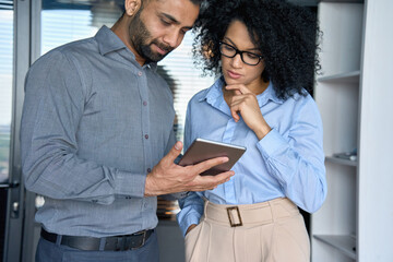 Focused Indian latin male ceo sales agent and female African American financial customer discussing financial business project looking at tablet device standing in modern corporate office.