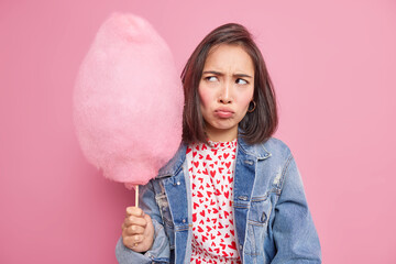 Dissatisifed sulking Asian teenage girl holds sweet cotton candy on stick doesnt like something wears denim jacket poses against pink background. Portrait of lovely female model with tasty dessert