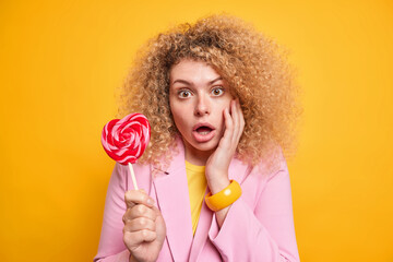 Portrait of curly haired woman looks amazed at camera keeps mouth opened keeps hand on face holds delicious candy dressed formally isolated over yellow background. Amazed female model with candy