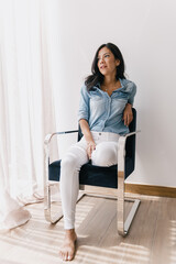 Portrait of Beautiful Hispanic or Latina Woman wearing white pants and a denim shirt sitting on a chair and looking at her window