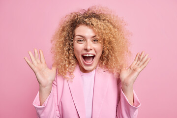 Excited curly haired European woman raises palms exclaims loudly keeps mouth opened reacts on awesome news wears formal jacket isolated over pink background. Positive human emotions concept.