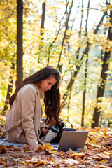 A brunette woman is sitting in an autumn park with a laptop and a cup of coffee. Student. Teacher. Online education.