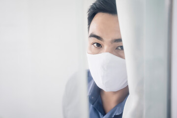 Asian man Self isolation quarantine lockdown social distancing at home looking out window peeking through curtain, wearing protection face mask staying safe from coronavirus covid-19 world pandemic