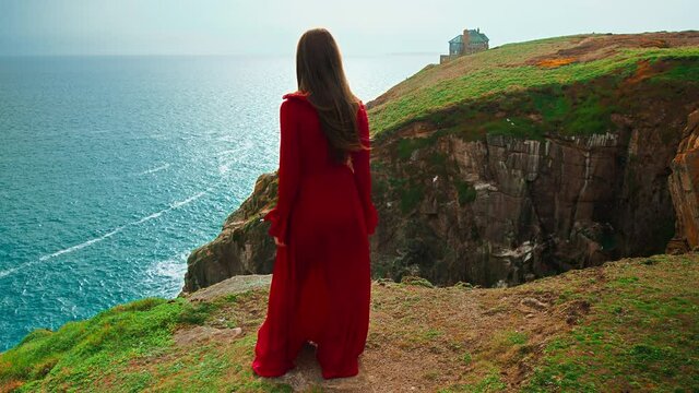 A beautiful girl in red dress standing at the edge of a granite rocky cliff. A young woman enjoying scenic views by a big seashore with a green grass on top and dream landscape views by Celtic Sea.