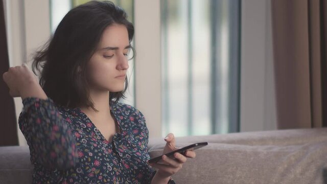 A girl sits on the couch, straightens her hair with her hand, uses her mobile phone, surfs the Internet