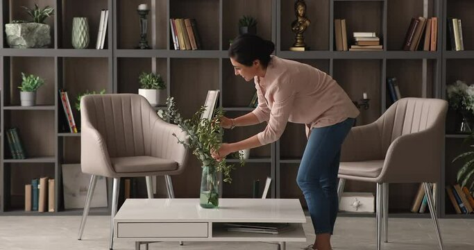 Young Indian woman bent over coffee table straighten bouquet in vase. Professional female decorator improve styling living room interior with flower arrangement. Housewife and housework chores concept