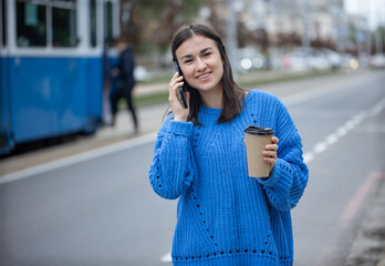 Stylish young girl speaks on the phone and holds coffee to go in her hand.