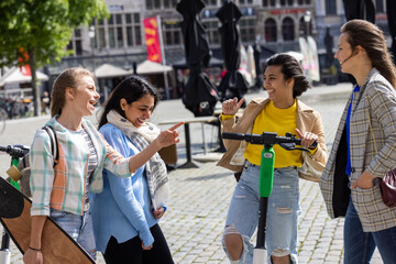 Lifestyle portrait of a diverse multiethnic group of four young girl friends on the electro scooter and skateboard having fun in a european city street at summer sunny day. Outdoor portrait of four
