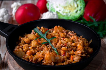 Homemade stew with vegetables, potato, onion, carrot, cabbage, pepper with tomato sauce, garlic and herbs in a frying dish on wooden table.