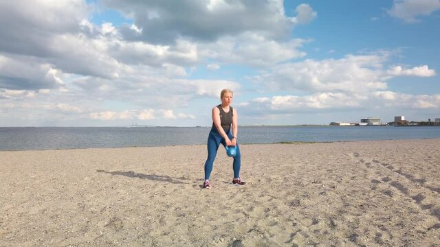 Young caucasian woman doing gym exercises and training on the beach in Amager, Copenhagen, Denmark. The girl is training with a kettle ball. It is a sunny spring evening. 3x times speeded up 4K clip