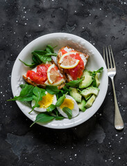 Bowl of green salad, avocado, boiled egg and sandwiches with rye bread, cream cheese and salmon on a dark background, top view. Delicious and healthy breakfast, brunch