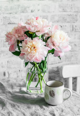 Pink peonies in a glass vase, tea cup on the table in a cozy room. Beauty home interior still life