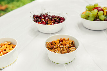 serving plate a mixture of nuts: almonds, hazelnuts, walnuts, ripe cherry berry, ripe juicy apples, grapes on light table in a open space green grass. healthy summer raw snacks. selective focus