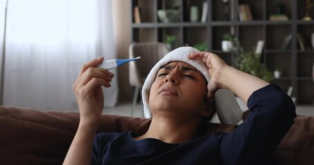 Indian frown unhealthy young woman lean on cushion of sofa having high fever put wet cloth on forehead holding electronic thermometer checks temperature feels unwell and weak, grippe symptoms concept
