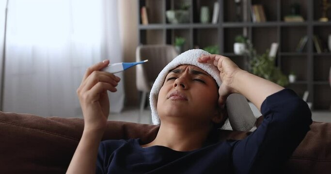 Indian frown unhealthy young woman lean on cushion of sofa having high fever put wet cloth on forehead holding electronic thermometer checks temperature feels unwell and weak, grippe symptoms concept