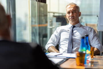 Two business men discussing their business together in a lobby of a hotel. They both wearing white shirt. The older one also wear black jacket. The scene outside is cityscape.