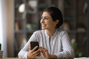 Dreamer with phone. Motivated indian female worker student sit at table look away hold cell think...