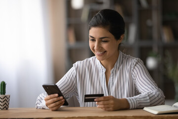 Confident ethnic female student pay tuition fee remotely using mobile phone bank app. Smiling young indian lady sit at office table use credit card to make online money transfer buy goods at internet - obrazy, fototapety, plakaty