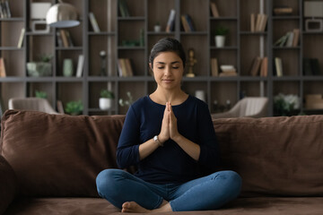 Relieving stress. Relaxed millennial indian woman sit on sofa alone in lotus position clasp hands in namaste. Serene young lady meditate at home breath deep with closed eyes feel harmony peace of mind